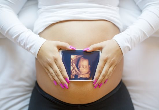 Close-up of an pregnant woman holding ultrasound photograph of a baby in front of the swollen bare tummy. Laying in the bed.